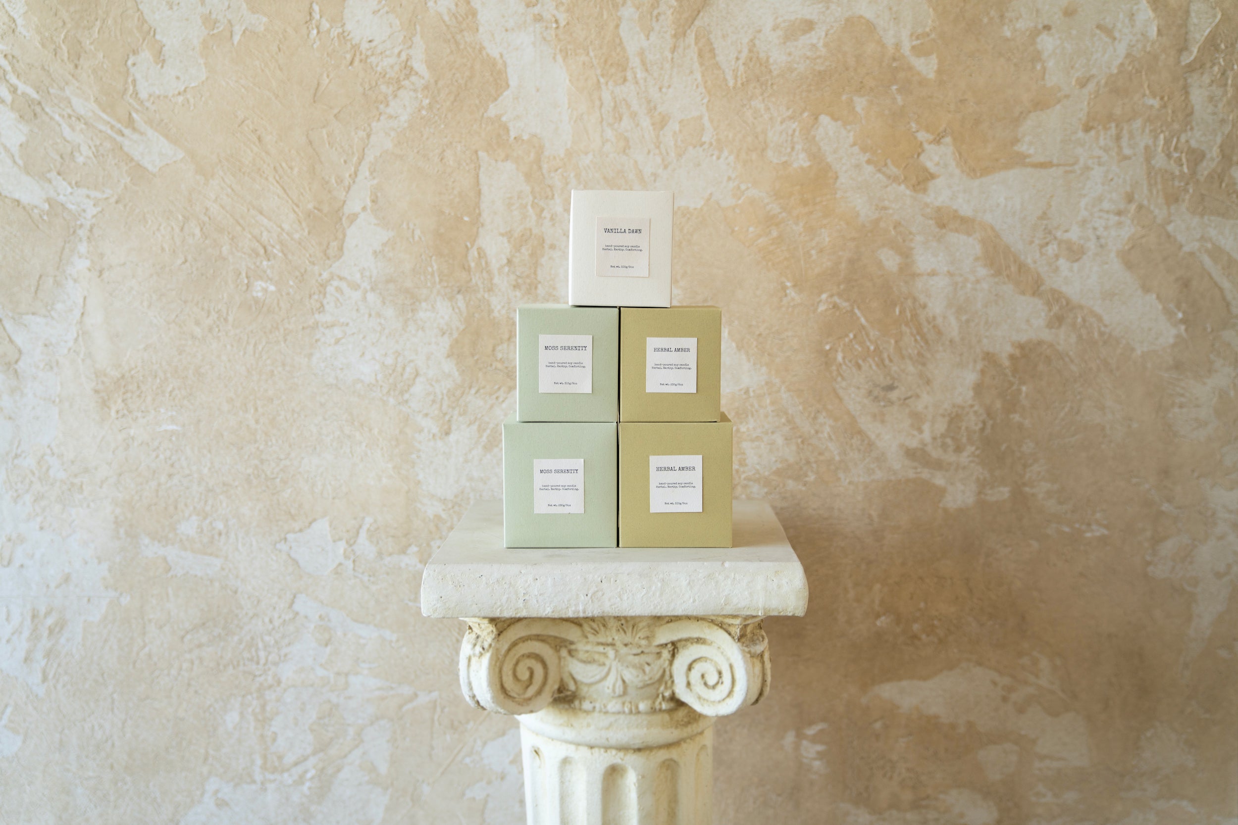 Stack of Enclify's minimalist candle packaging boxes, showcasing Herbal Amber, Moss Serenity, and Vanilla Dusk in a cohesive, earthy palette