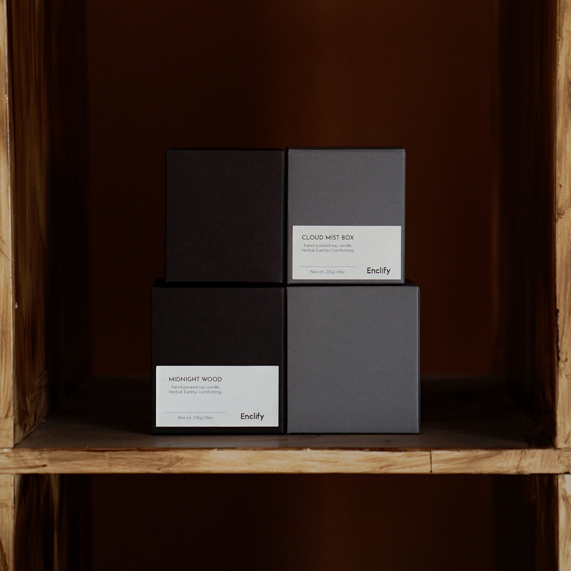 Dark and light grey Enclify candle boxes, including Midnight Wood, on a wooden shelf.